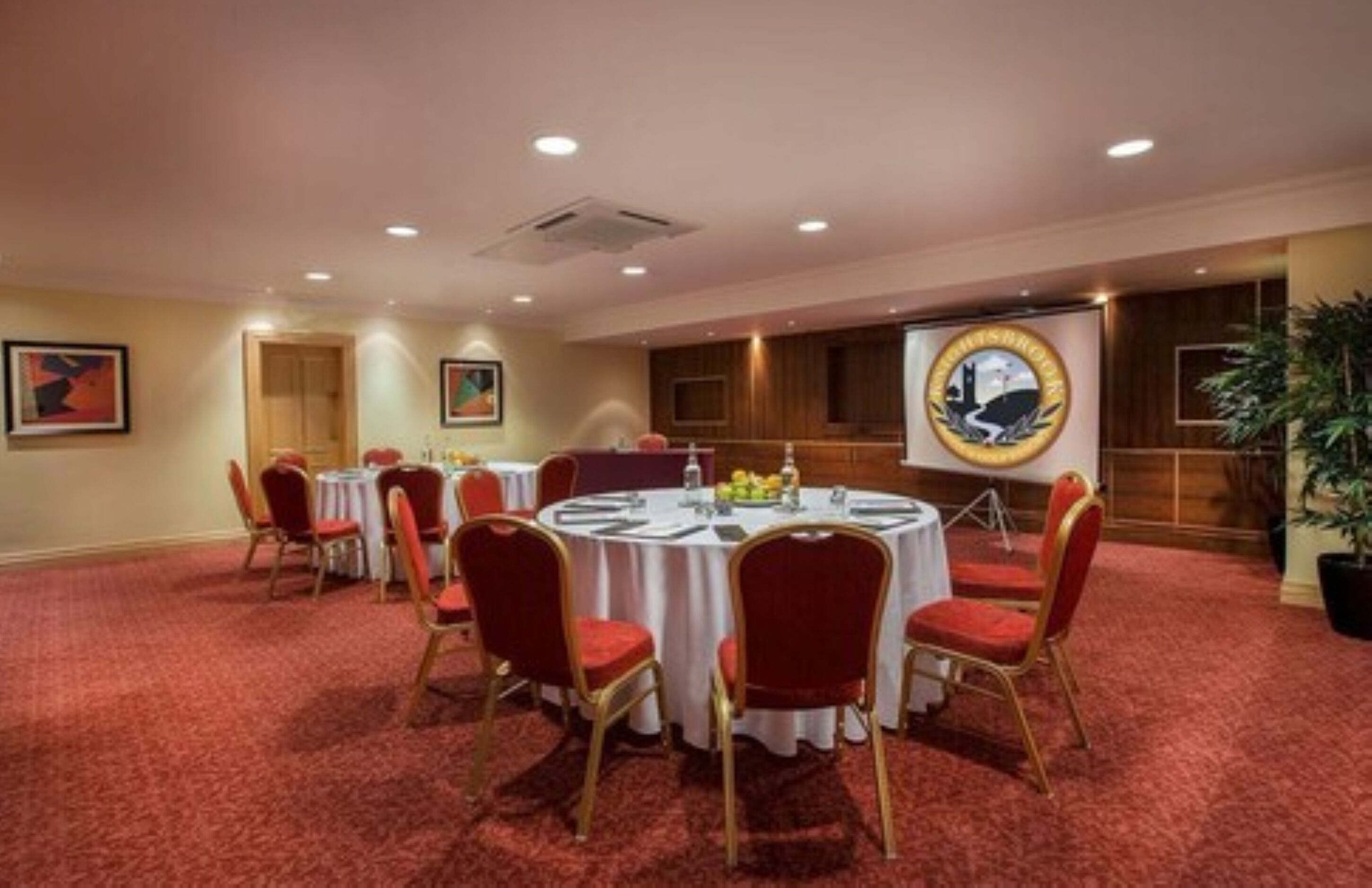 A conference room with round tables, red chairs, and a projector screen displaying a logo.