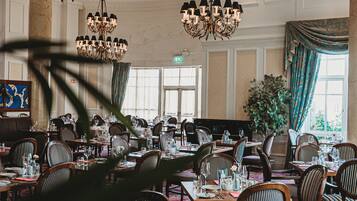 Un comedor amplio con varias mesas y sillas, candelabros elegantes y ventanas grandes que dejan entrar luz natural.