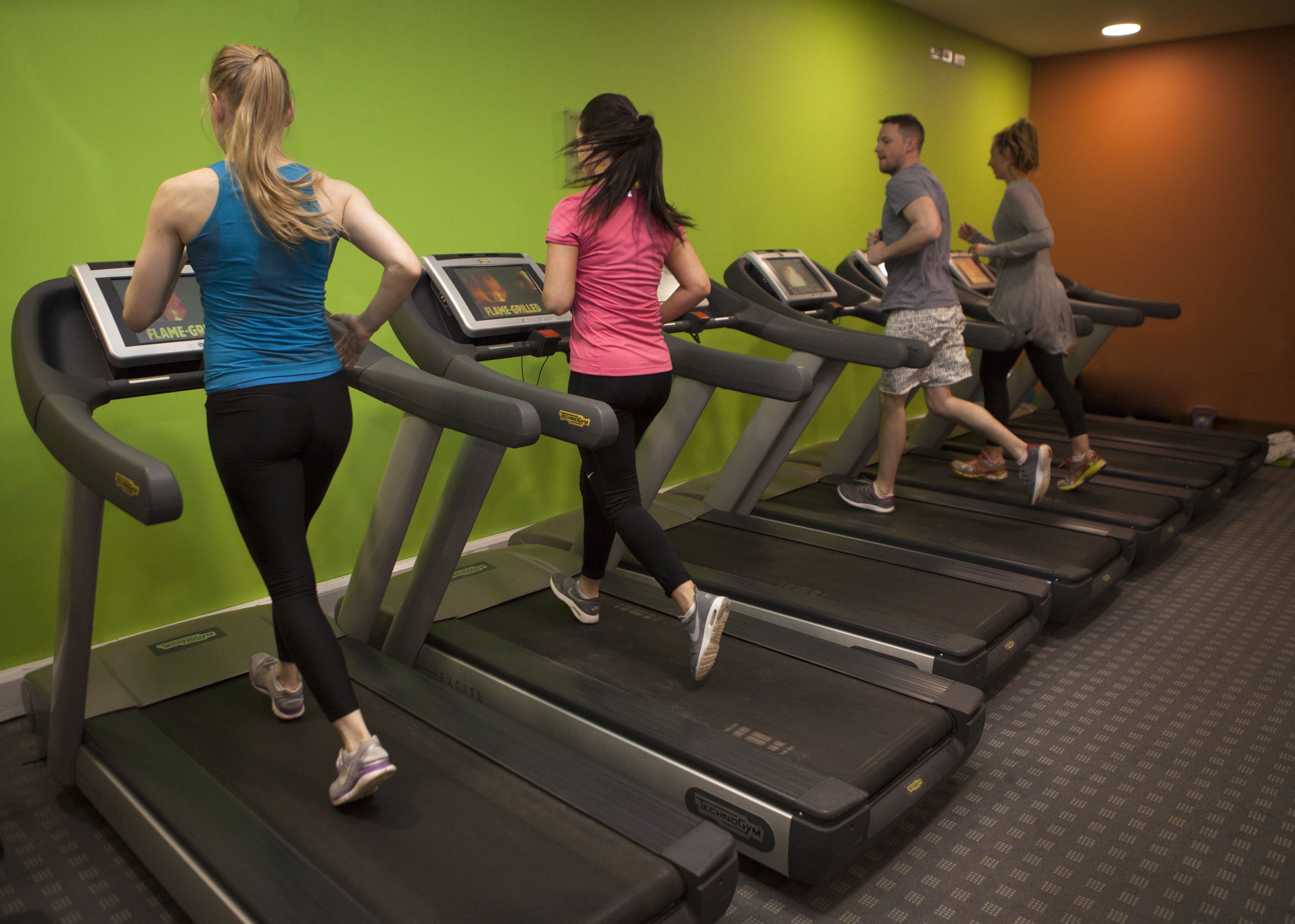 En grupp människor som använder löpband i ett gym med gröna väggar och ett mönstrat golv.