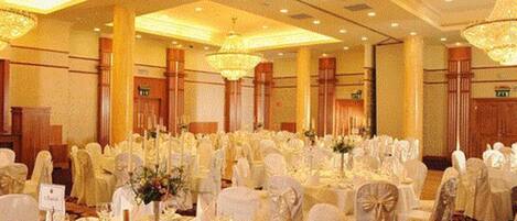 Un salón de banquetes con mesas redondas, manteles blancos y sillas decoradas con lazos.