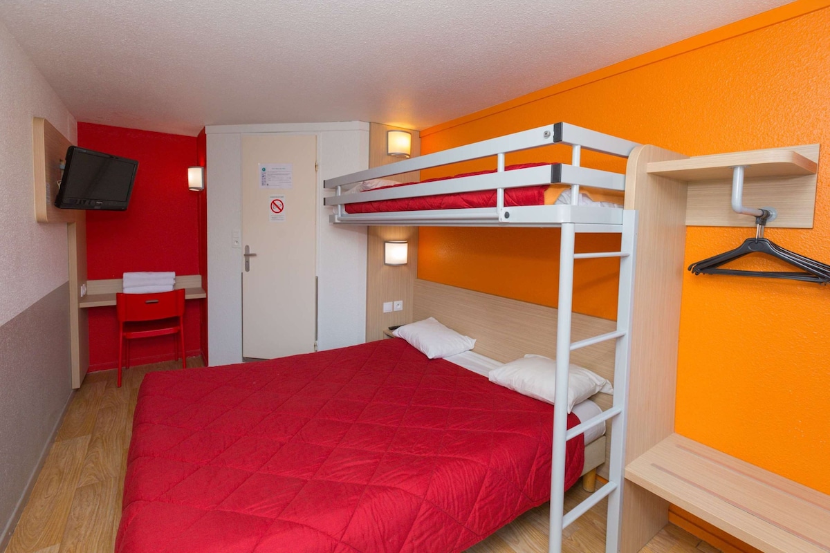 Habitación con literas, una colcha roja, un escritorio con silla, un televisor montado en la pared y una puerta con un cartel.