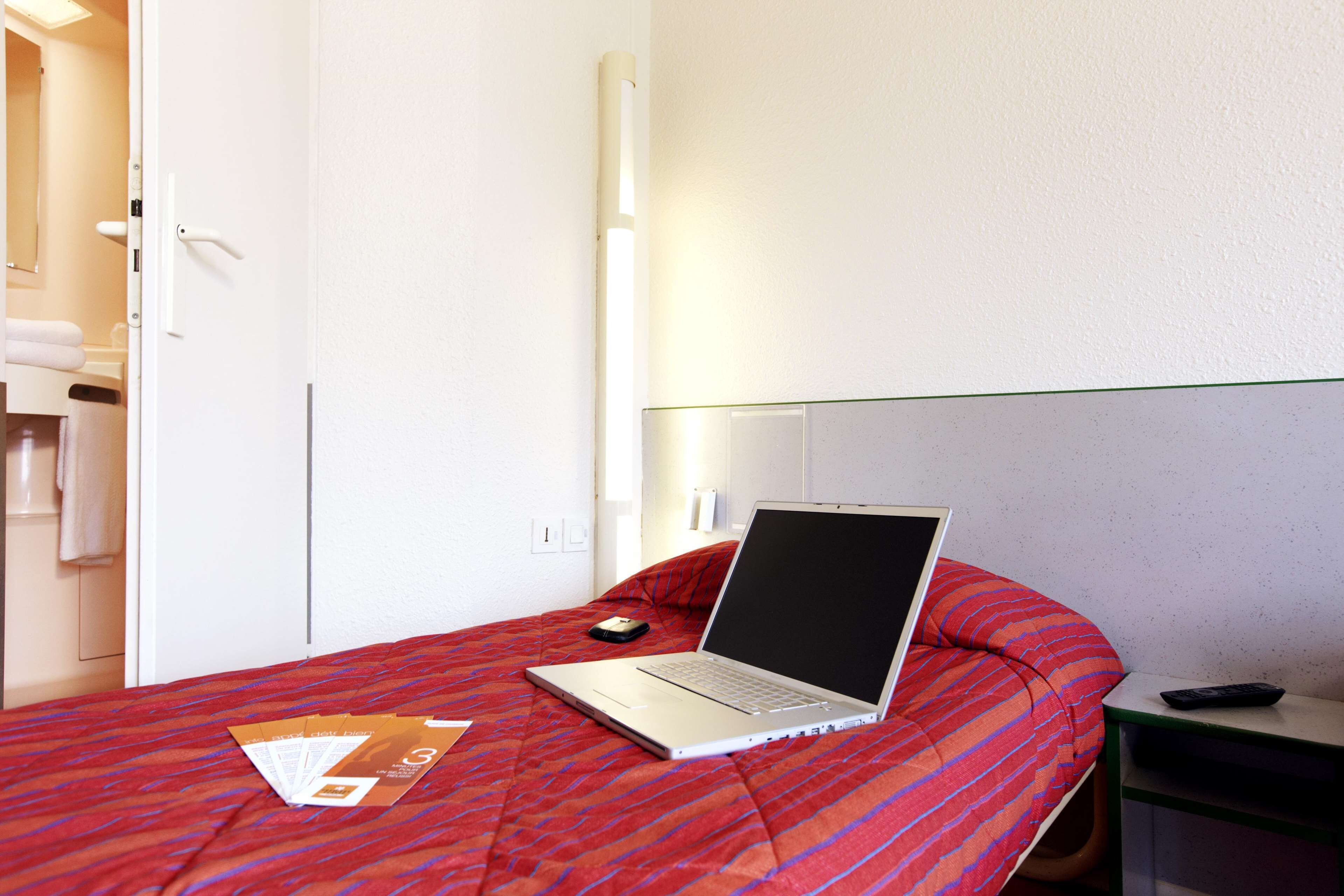 A hotel room with a bed, a laptop, a remote control, and a door to the bathroom.