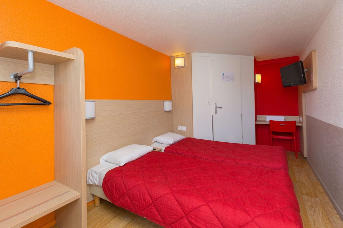 Habitación de hotel con una cama roja, un armario de madera, un escritorio con silla y un televisor en la pared.