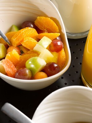 Un tazón de ensalada de frutas mixtas con una cuchara, sobre una superficie negra junto a un tazón blanco y un vaso de jugo de naranja.