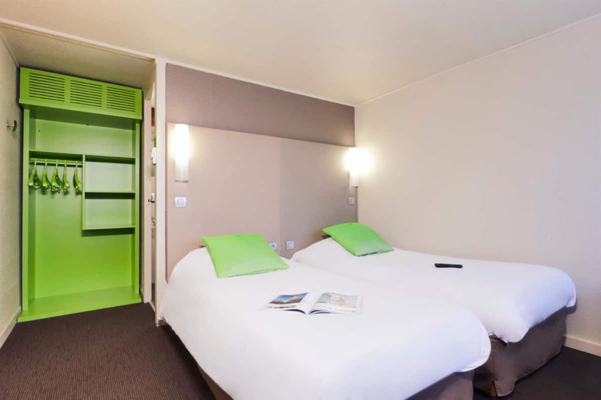 Habitación de hotel con dos camas, almohadas verdes, una revista sobre la cama de la izquierda y un armario verde con ropa colgada.