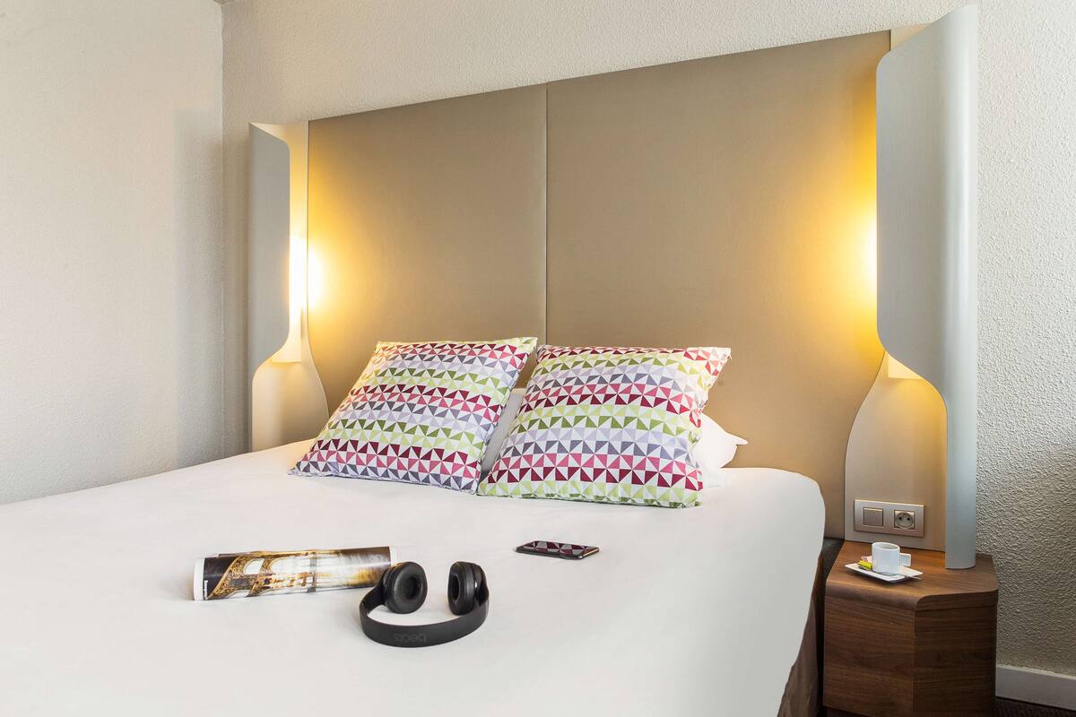 Una cama bien hecha con almohadillas estampadas, una mesita de noche con lámpara, una revista, audífonos y un teléfono inteligente sobre la cama.