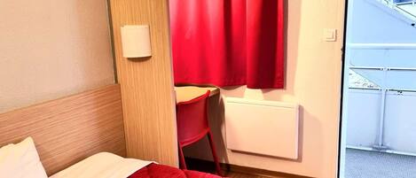 Une chambre d’hôtel avec une couvre-lit rouge matelassé, une tête de lit en bois, une chaise rouge et une fenêtre donnant sur un immeuble et de la verdure.