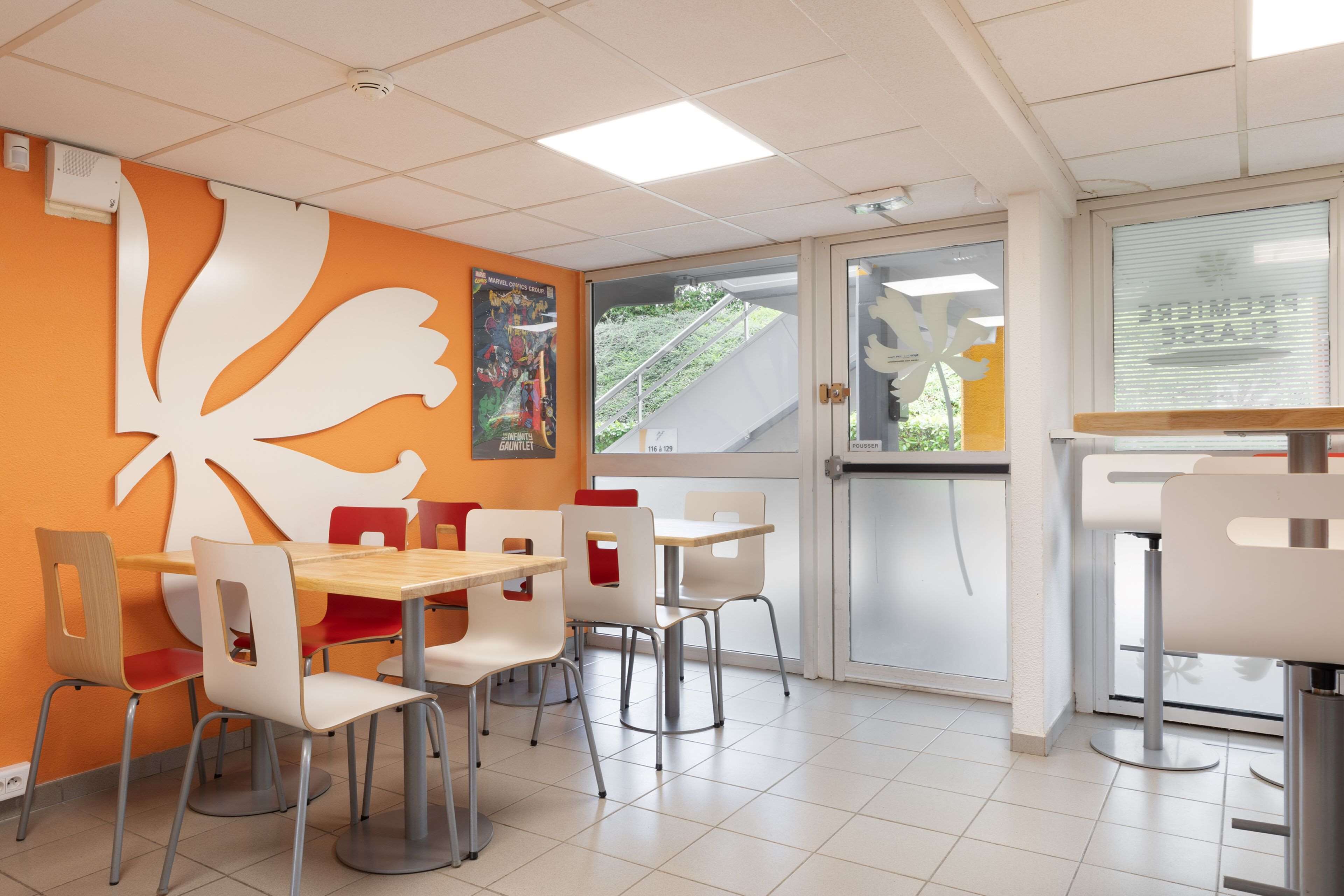 A modern restaurant interior with orange walls, white and red chairs, and a large decorative wall piece.