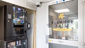 A vending machine with a selection of beverages and snacks, a surveillance camera, and a glass door with a sign indicating the entrance to Premiere Classe Hotels.