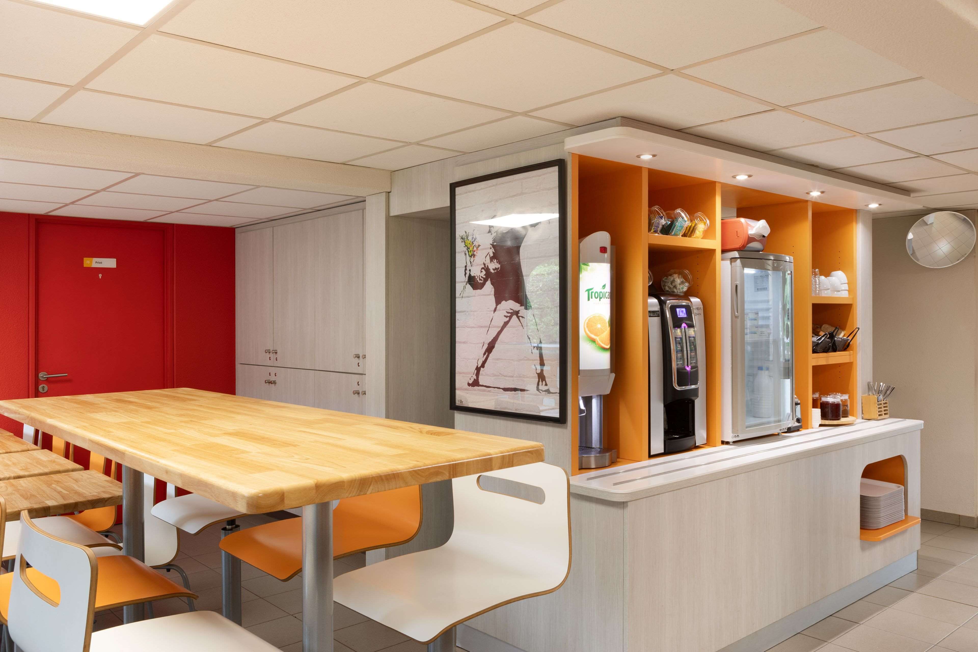 A modern cafe with wooden tables and white chairs, a counter with a beverage dispenser, and a red door in the background.