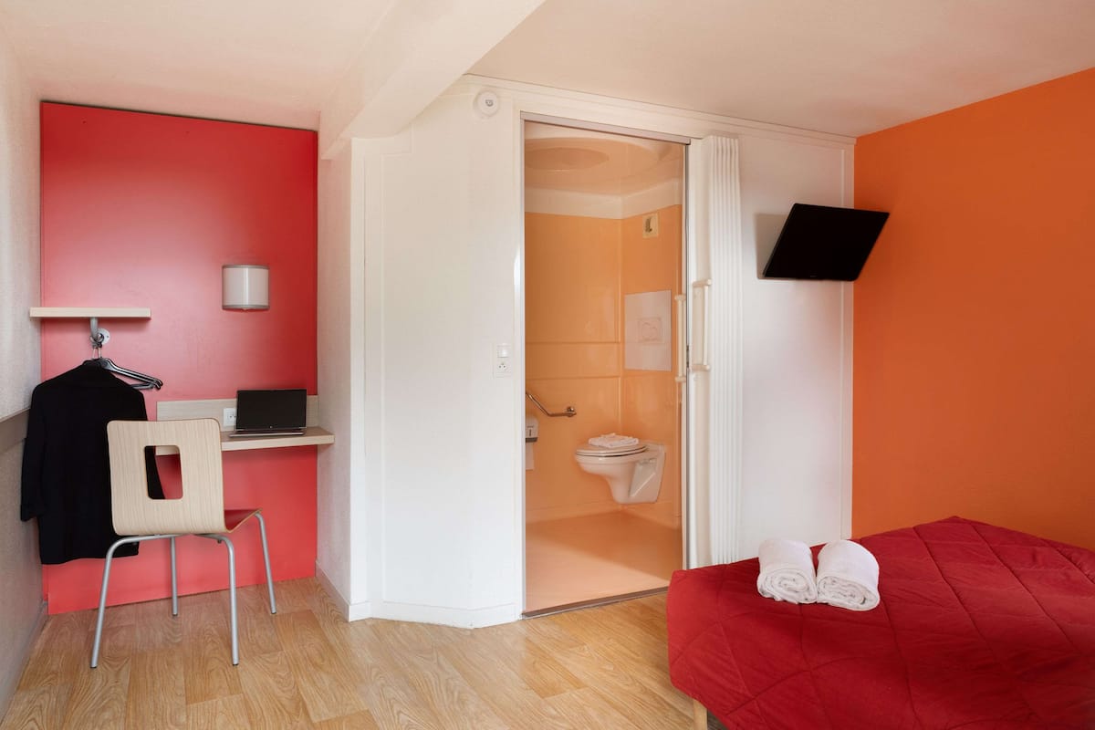 Ein Zimmer mit einem roten Bett, einem Schreibtisch mit einem Laptop, einem Stuhl und einem Duschbereich.