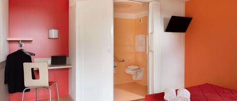 Una habitación con una cama roja, un escritorio con una computadora portátil, una silla y una zona de ducha.