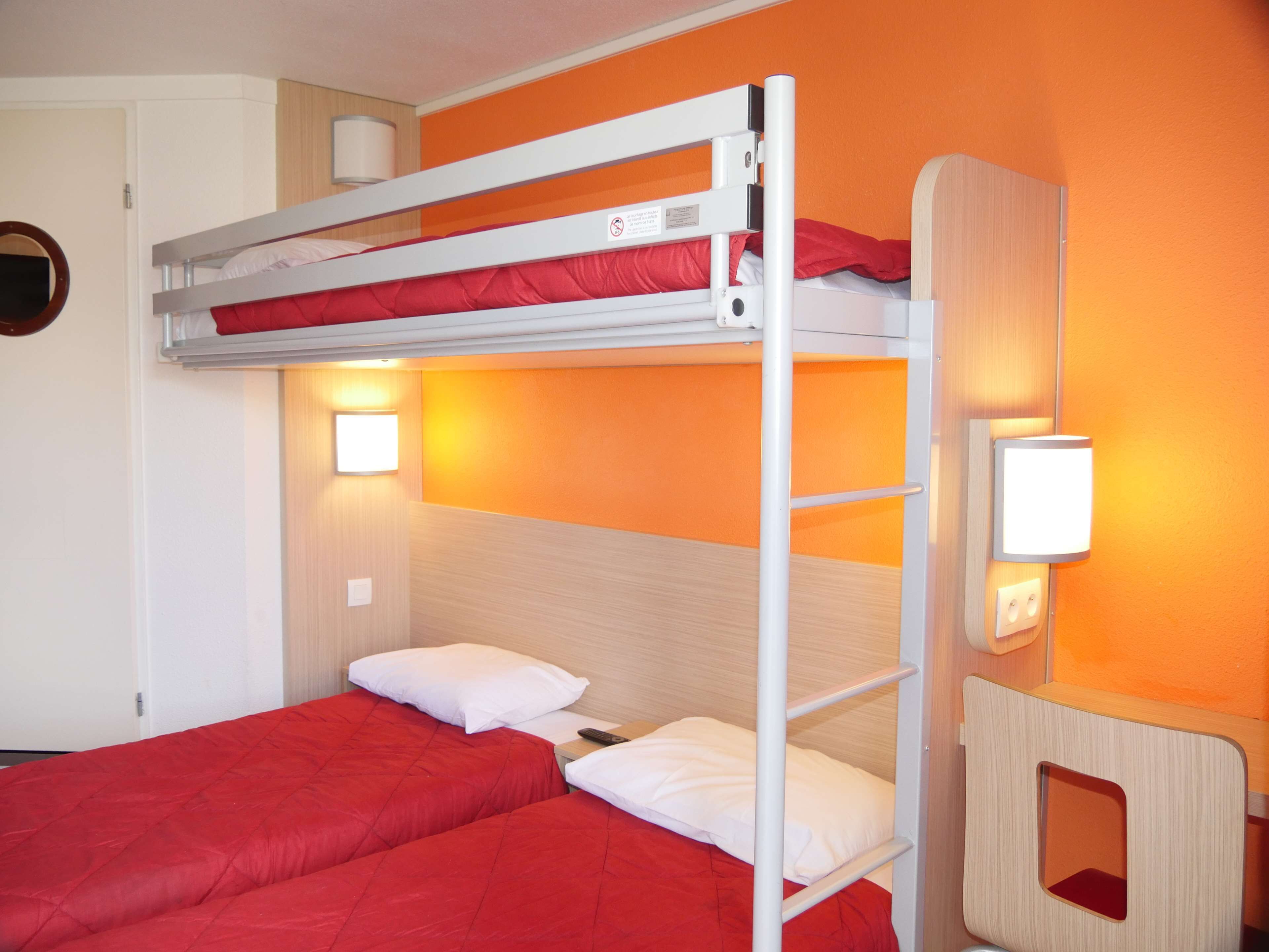 A bunk bed room with two beds, each with red bedding and white pillows. There are two wall-mounted lights and a wooden panel with a cut-out window on the right side.