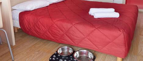 A single bed with a red quilt, two white pillows, and two metal pet bowls on a black mat with paw prints.