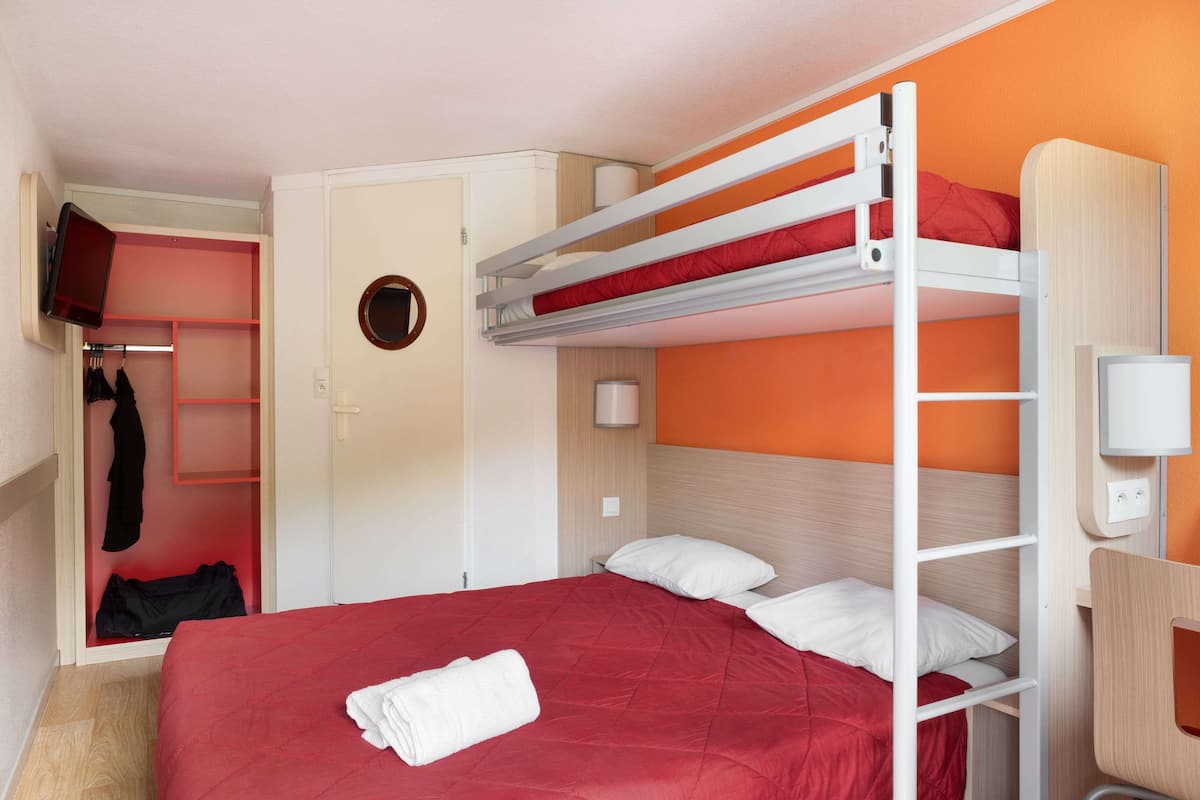 Ein Etagenbett mit roter Bettdecke und weißen Kissen, ein weißer Kleiderschrank, ein an der Wand befestigtes Fernsehgerät und ein kleiner runder Spiegel.