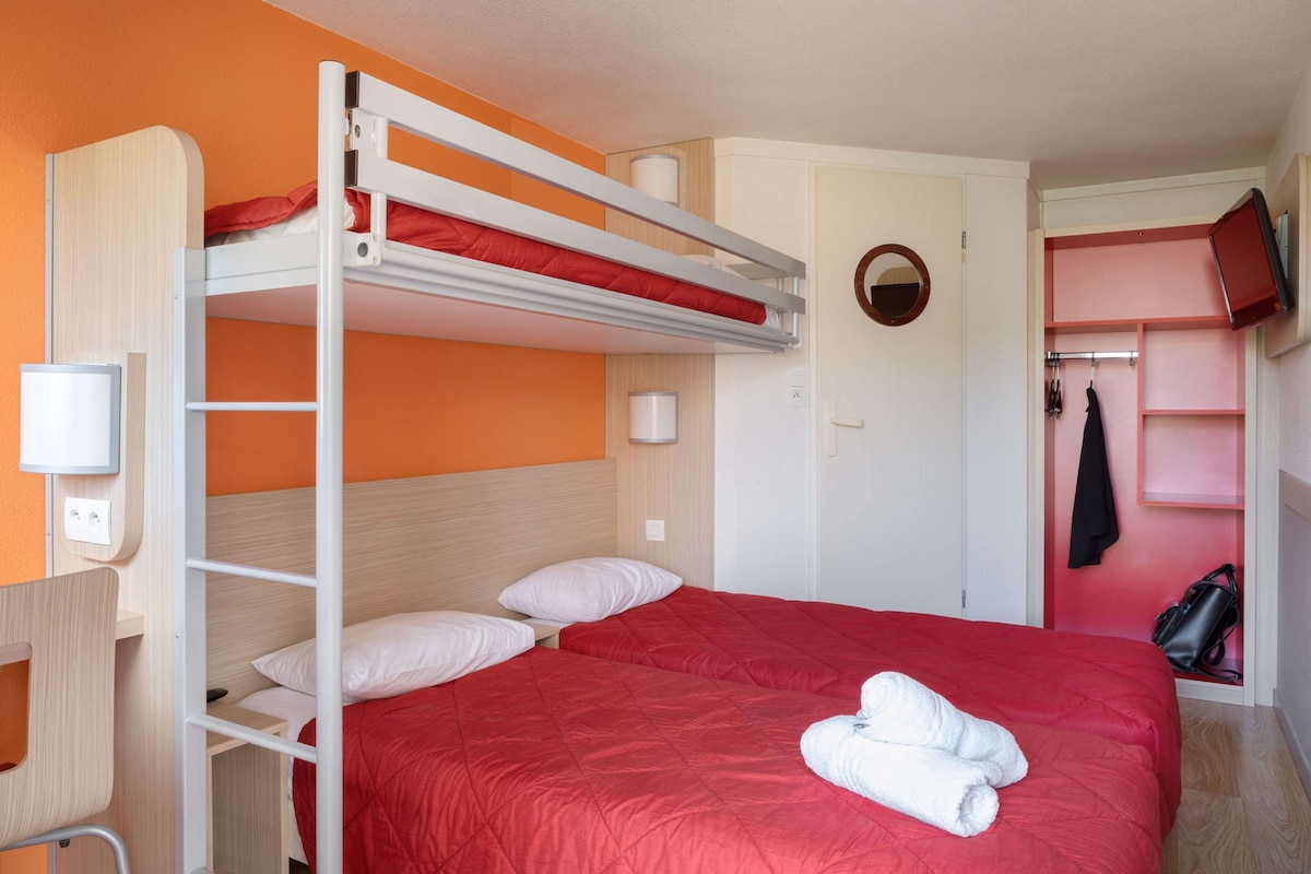 Una cama litera con un edredón rojo y almohadas blancas, un escritorio con una silla, un televisor montado en la pared y un perchero con una chaqueta.