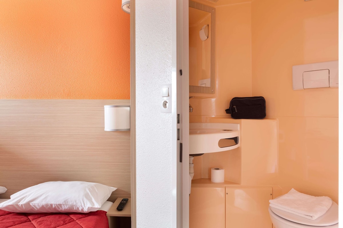 Ein Hotelzimmer mit einem Bett, einem Nachttisch, einer Fernbedienung, einem weißen Kopfkissen, einer roten Decke, einem Badezimmer mit Waschbecken, einem Spiegel, einem Handtuch und einer schwarzen Tasche.
