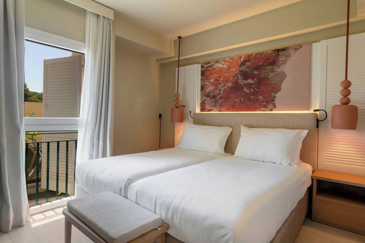 A double bed with white linens, a bedside table, a bench, a large abstract painting, and a balcony with white curtains.