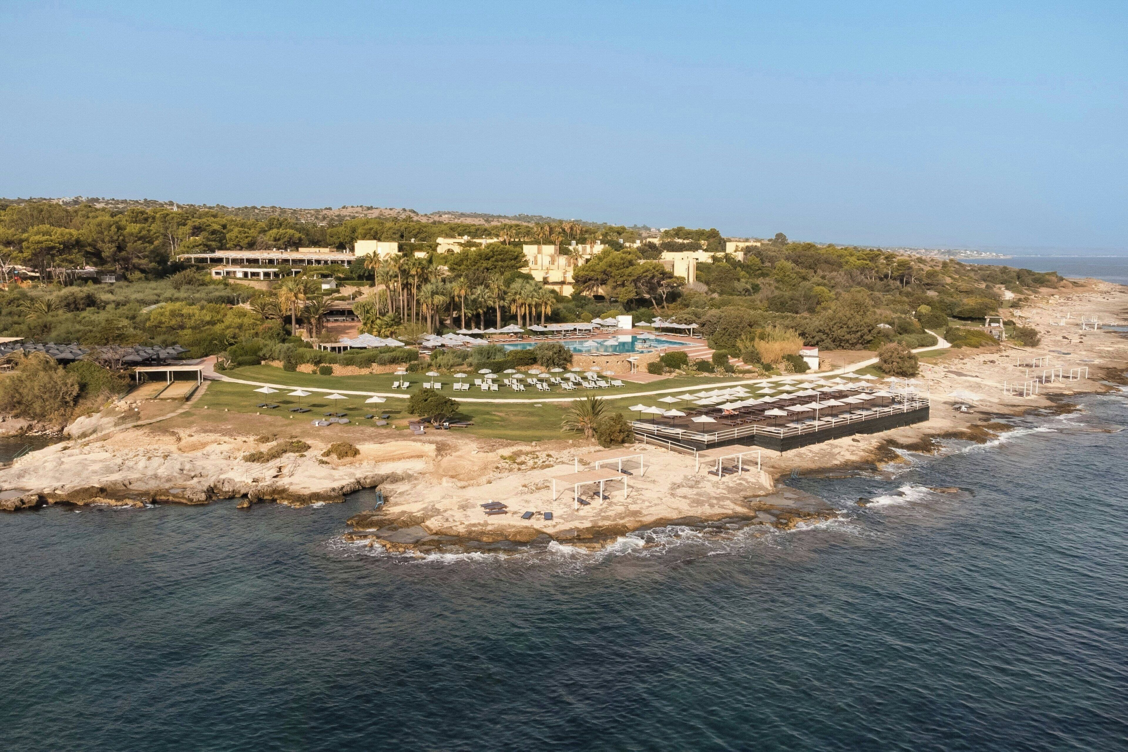 Vista aérea de un complejo turístico frente al mar, con varios edificios, una piscina y una zona de playa.