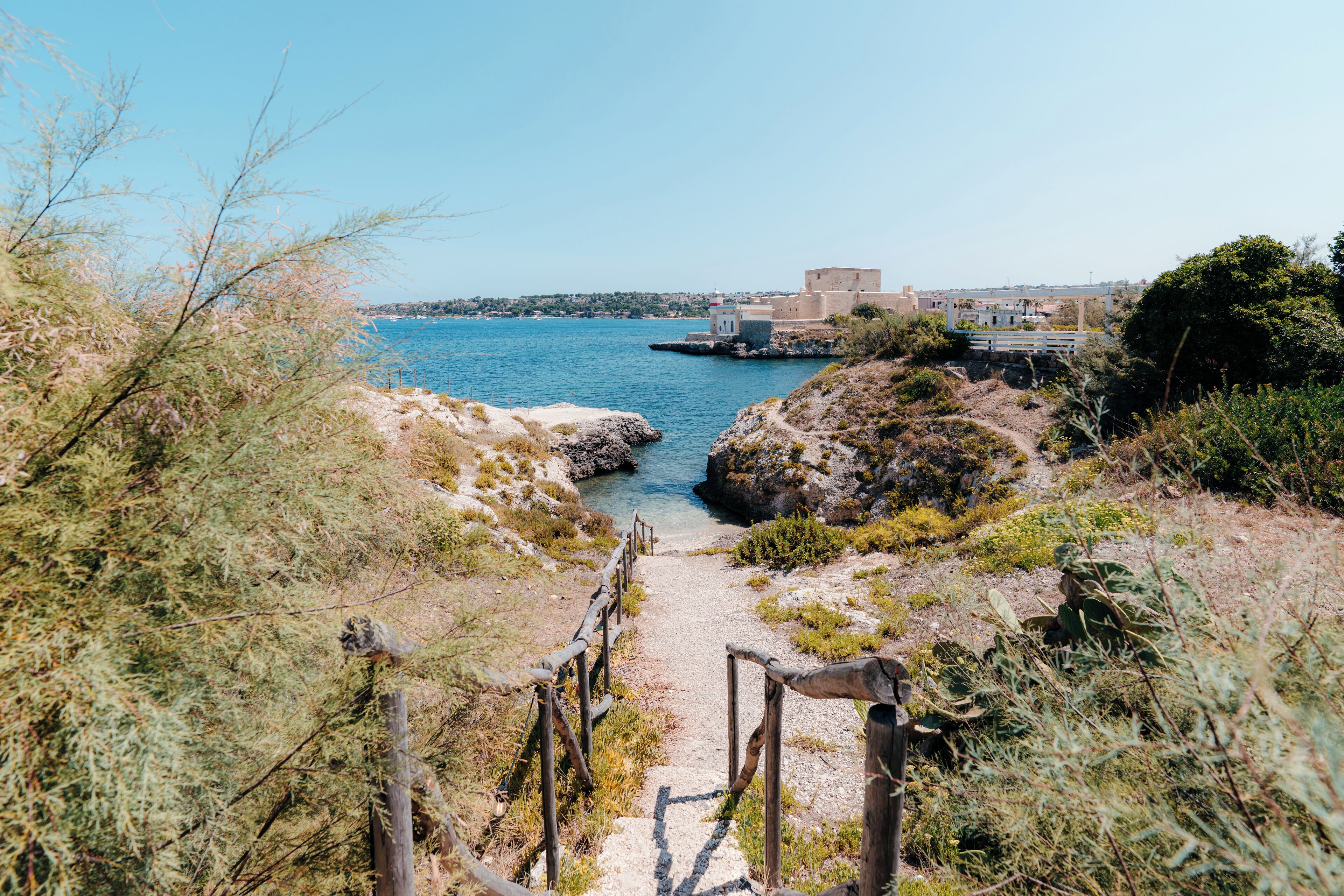 Un sentier côtier qui mène à une plage rocheuse, offrant une vue dégagée sur la mer et les bâtiments situés au loin.