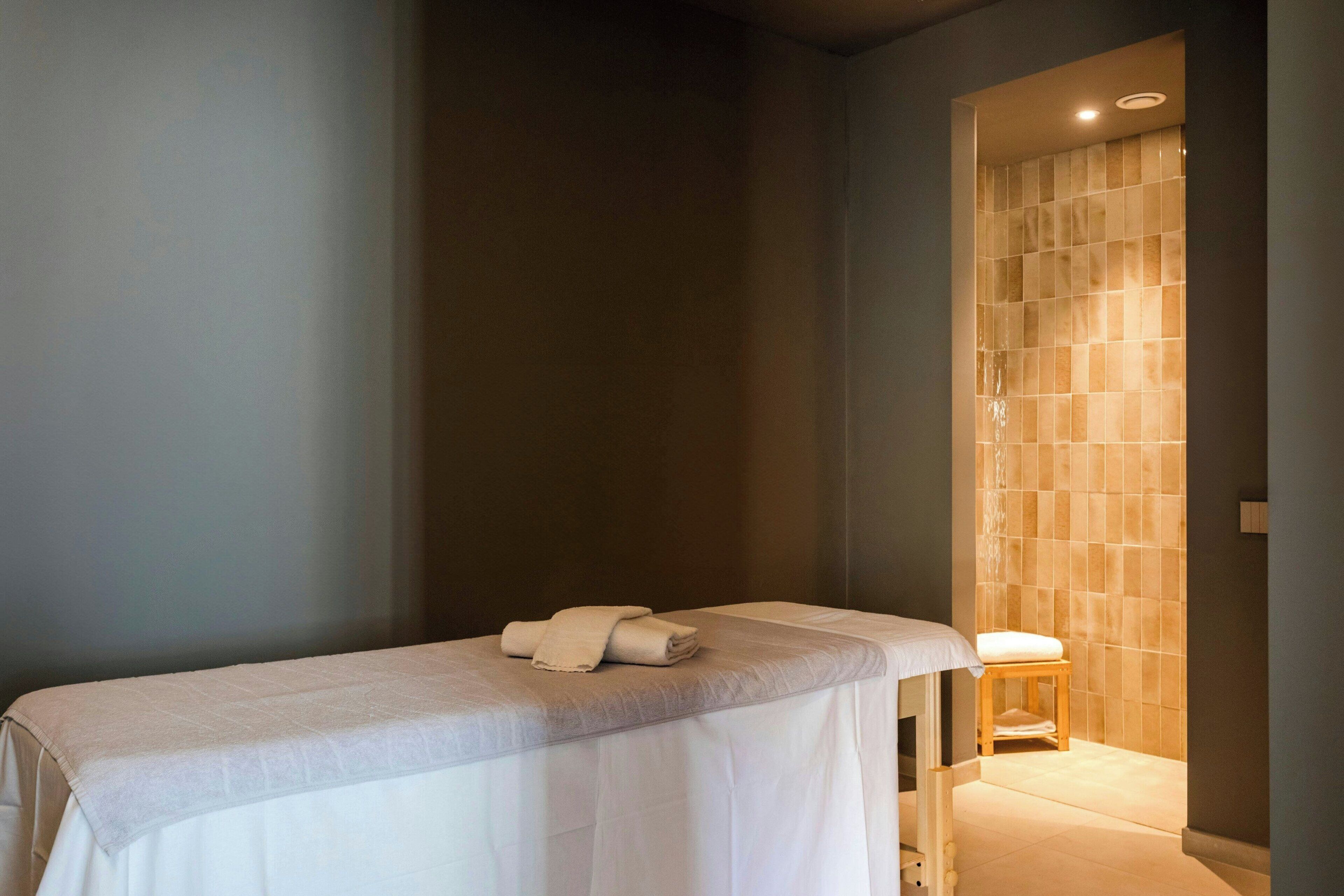 A spa room with a massage table, a wooden stool, and a tiled wall.