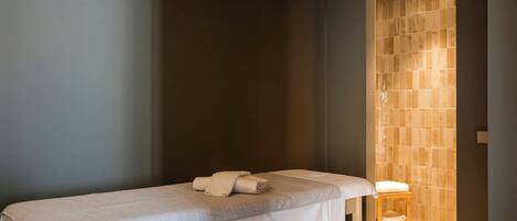 A spa room with a massage table, a wooden stool, and a tiled wall.