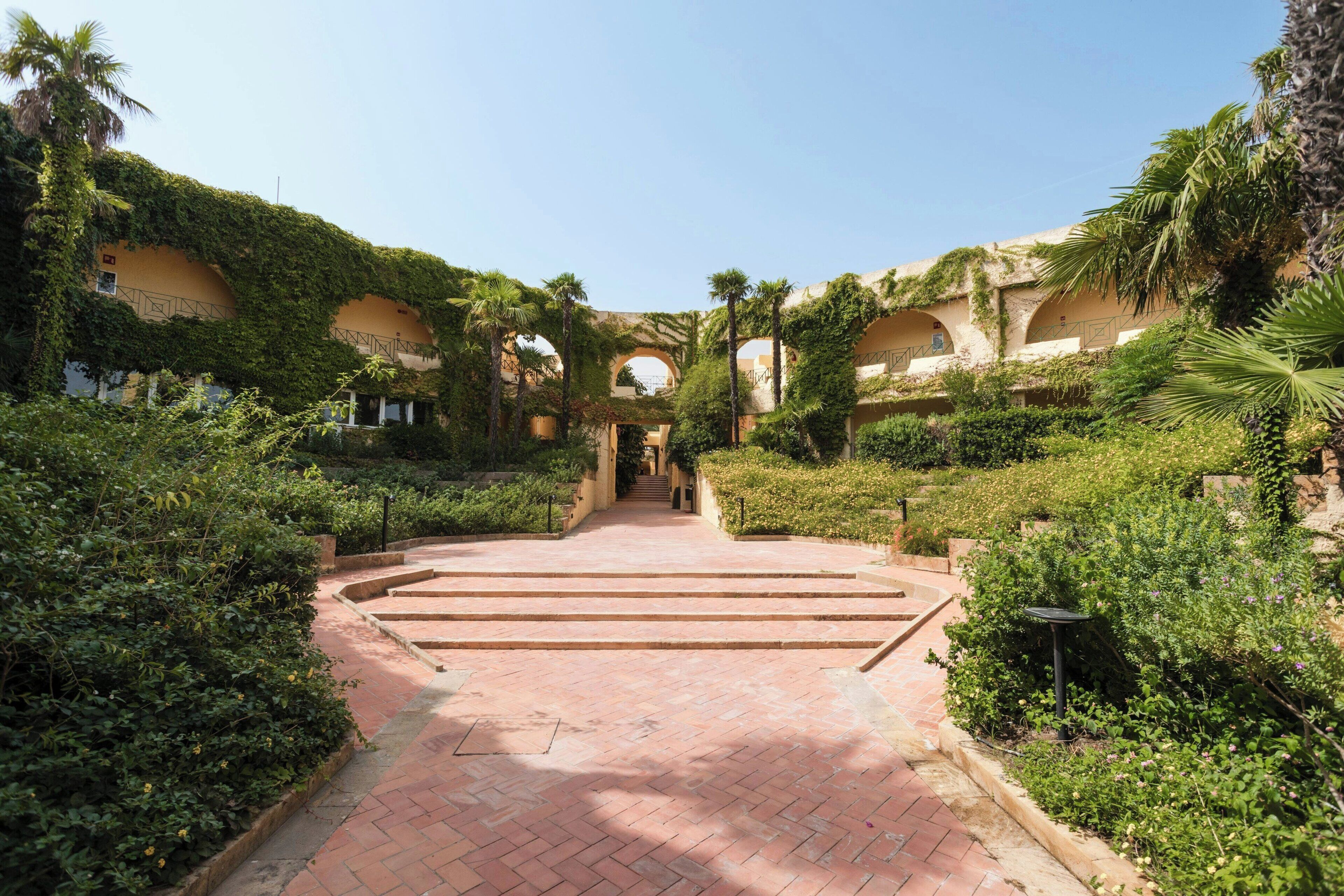 A courtyard with a brick pathway leading to a central archway, surrounded by lush greenery and palm trees.