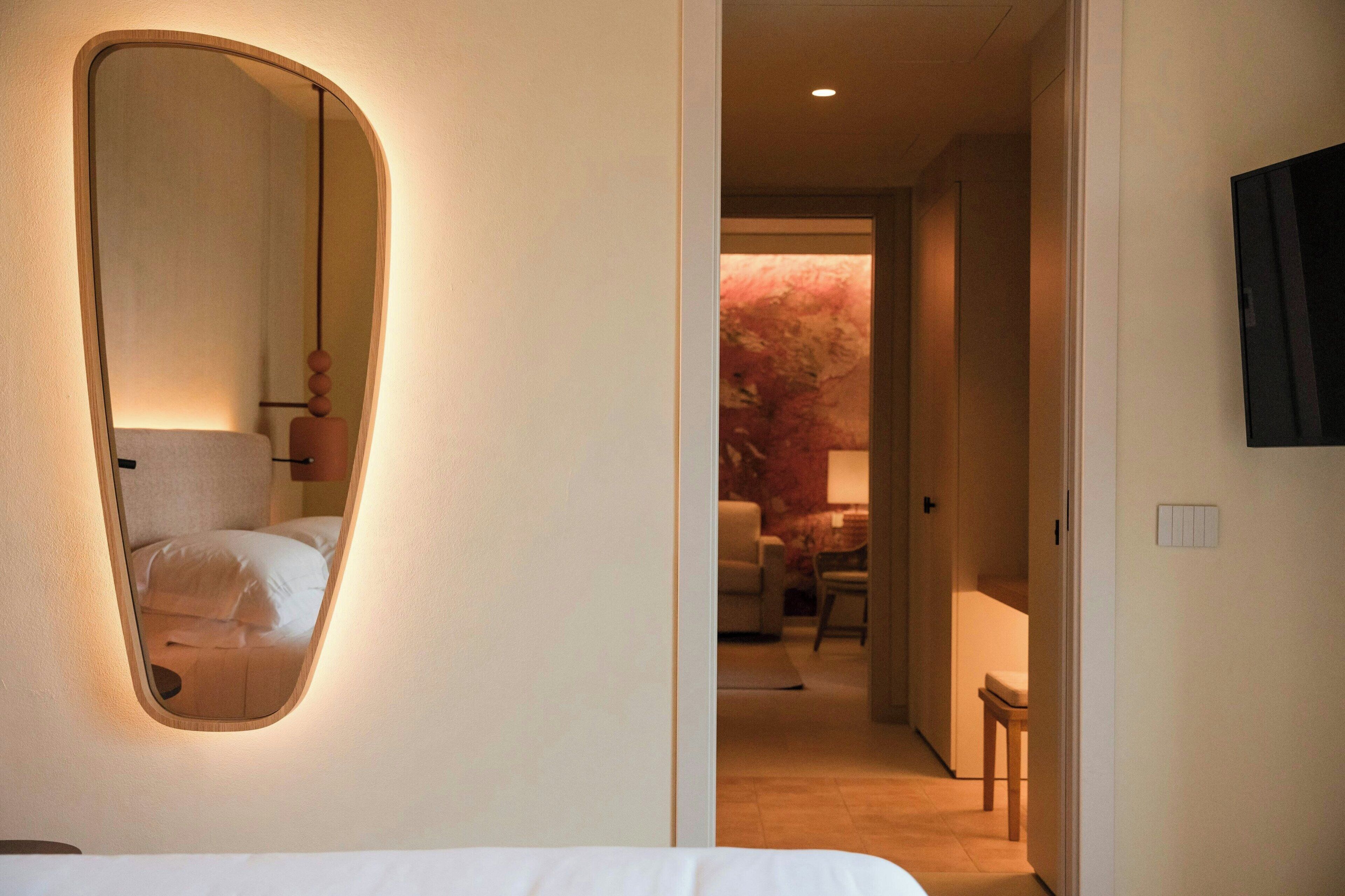 Habitación de hotel con un espejo grande, una cama, un televisor montado en la pared y vistas a un pasillo con una silla y una mesa.