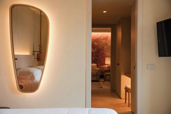 A hotel room with a large mirror, a bed, a TV mounted on the wall, and a view of a hallway with a chair and a table. - Mangia's Brucoli, Sicily, Autograph Collection (Augusta)