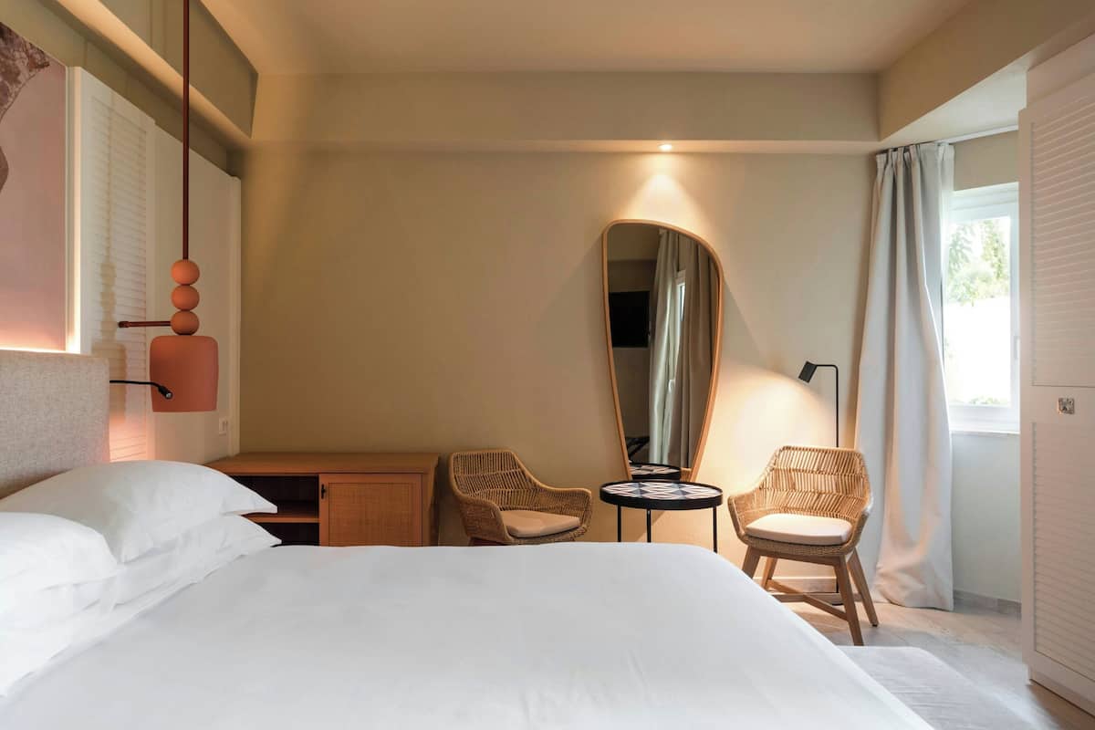 A hotel room with a large bed, two wicker chairs, a small round table, a mirror, and a desk with a lamp.