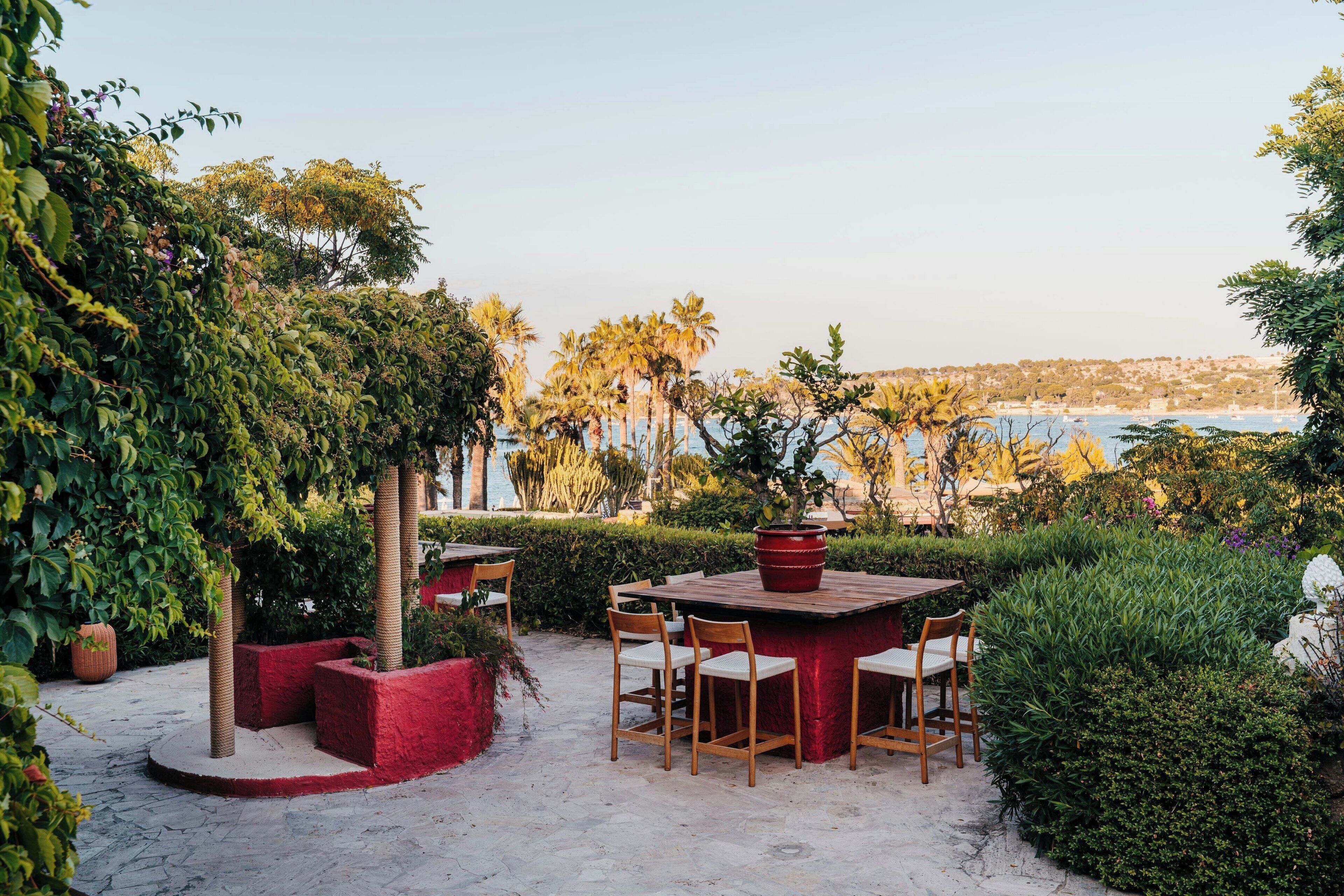 An outdoor patio with a round table and chairs, surrounded by lush greenery and palm trees, overlooking a coastal area.