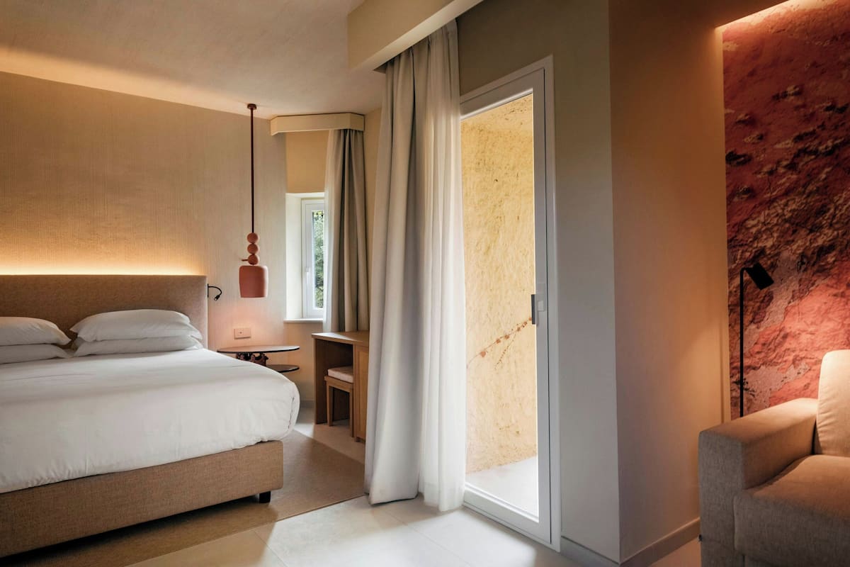 A modern hotel room with a large bed, a desk, and a chair. There is a sliding glass door leading to a balcony.