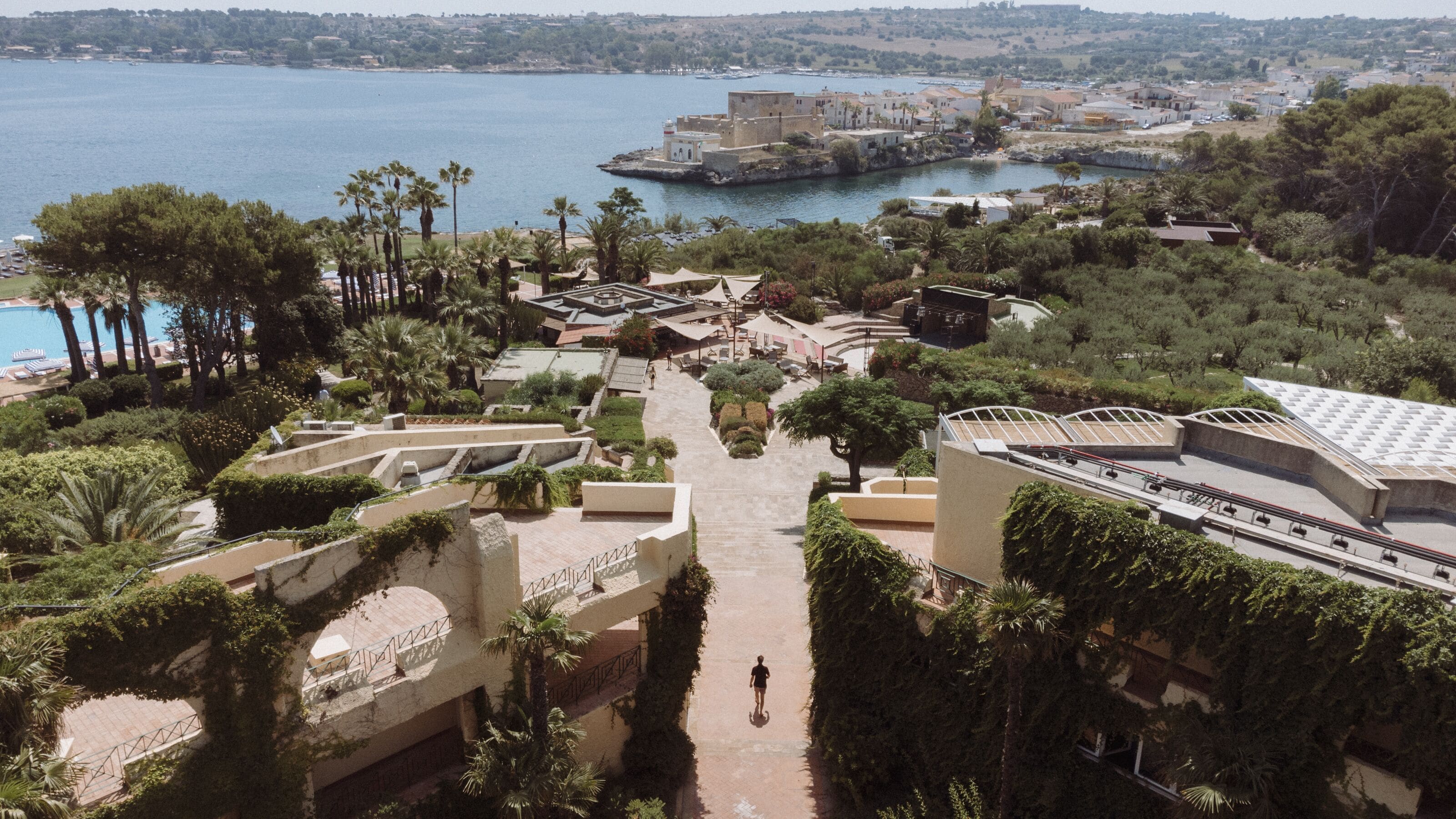 A coastal resort with a central courtyard, palm trees, and a view of the sea.