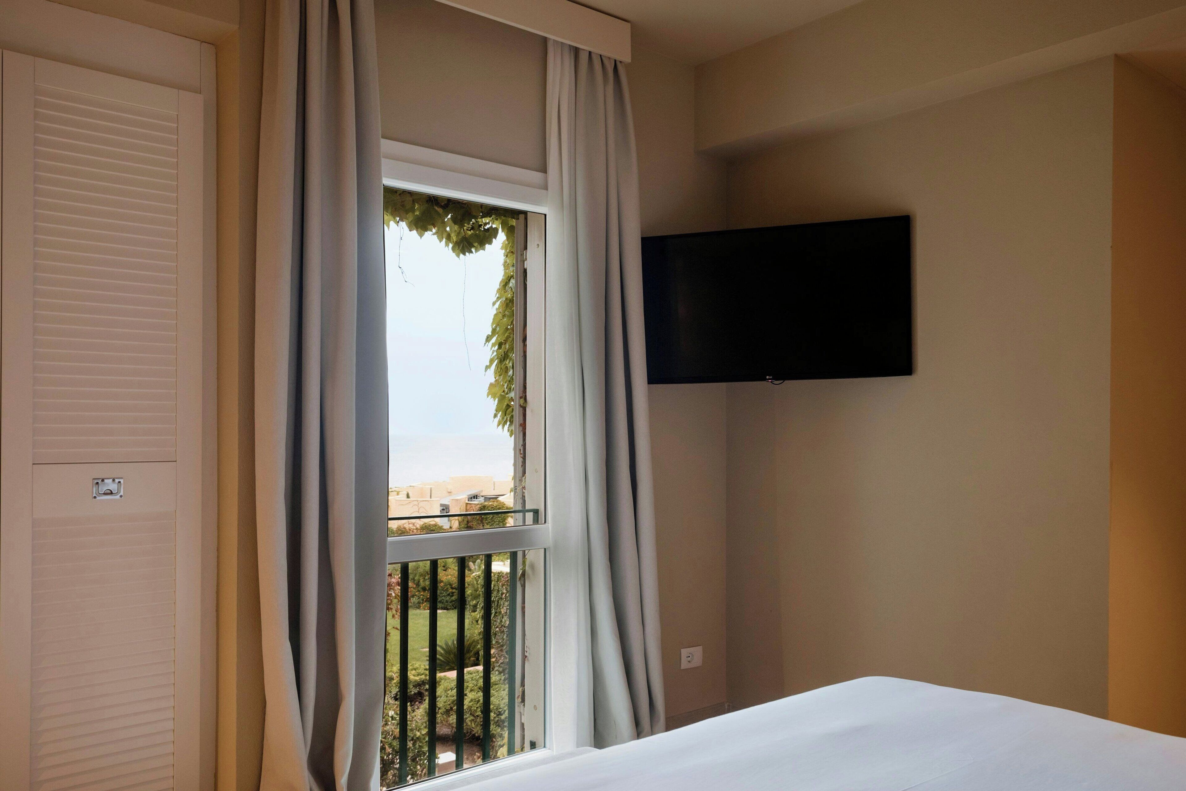 Ein Hotelzimmer mit einem Bett, einem an der Wand befestigten Fernseher und einem Fenster mit Vorhängen.