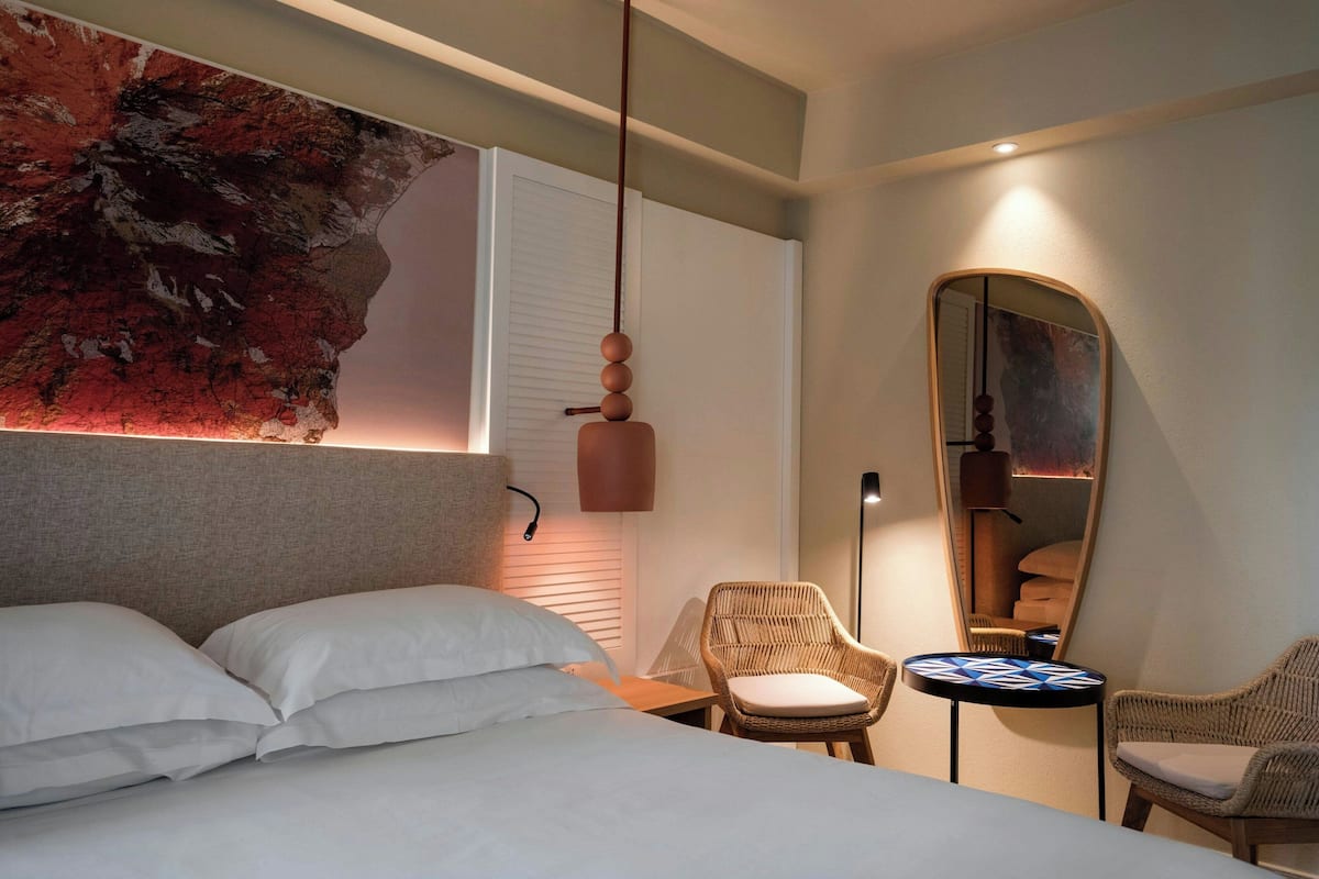 A modern hotel room with a large bed, a wall-mounted artwork, a round mirror, and two wicker chairs.