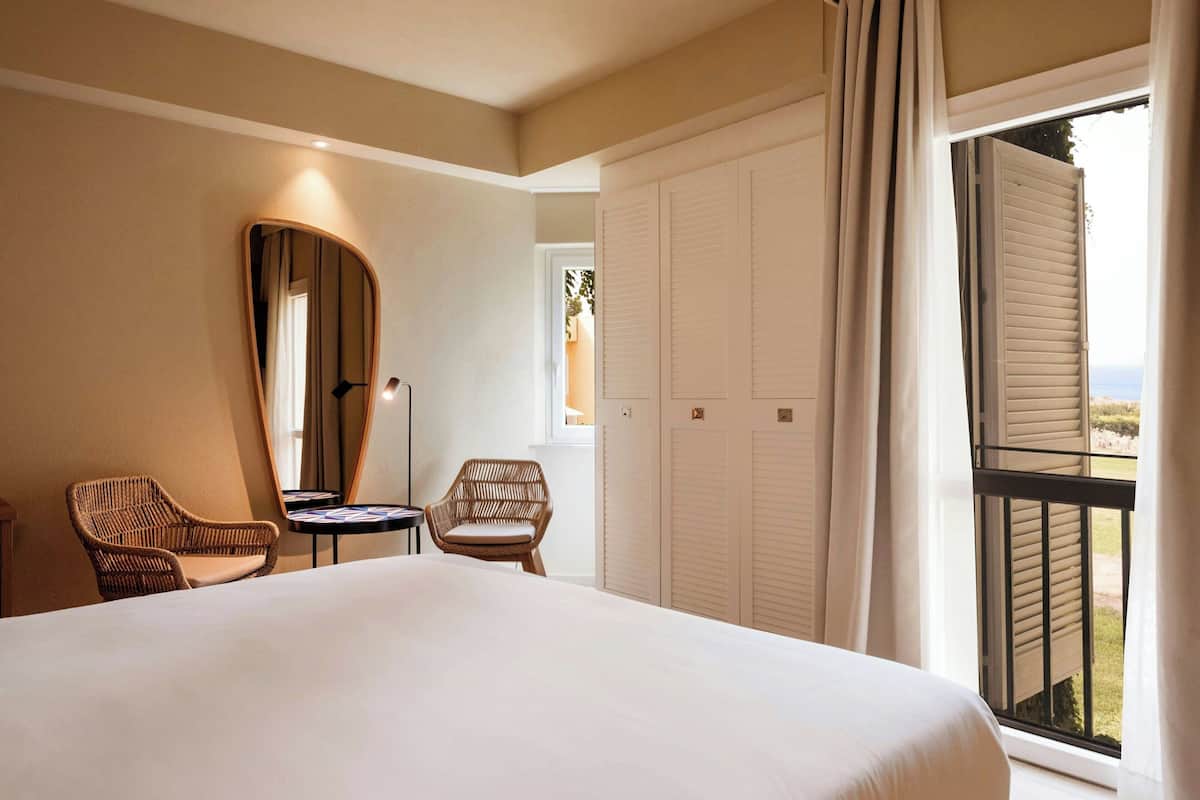 A hotel room with a large bed, two wicker chairs, a small table, a mirror, and a view of the outdoors through a window.
