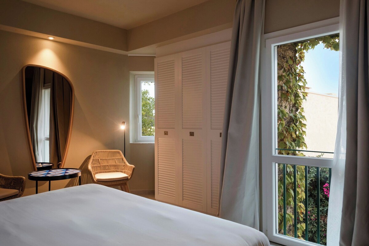 A hotel room with a bed, a round table with a board game, a wicker chair, a mirror, and a window with a view of greenery.