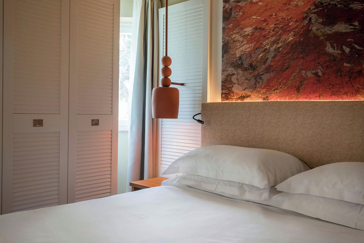 A neatly made bed with white linens, a headboard with a textured fabric, and a wall-mounted light fixture.
