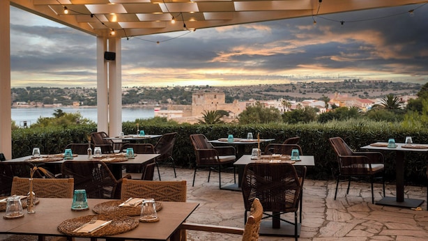 An outdoor dining area with tables and chairs, a view of a distant city and castle, and a pergola with string lights. - Mangia's Brucoli, Sicily, Autograph Collection (Augusta)