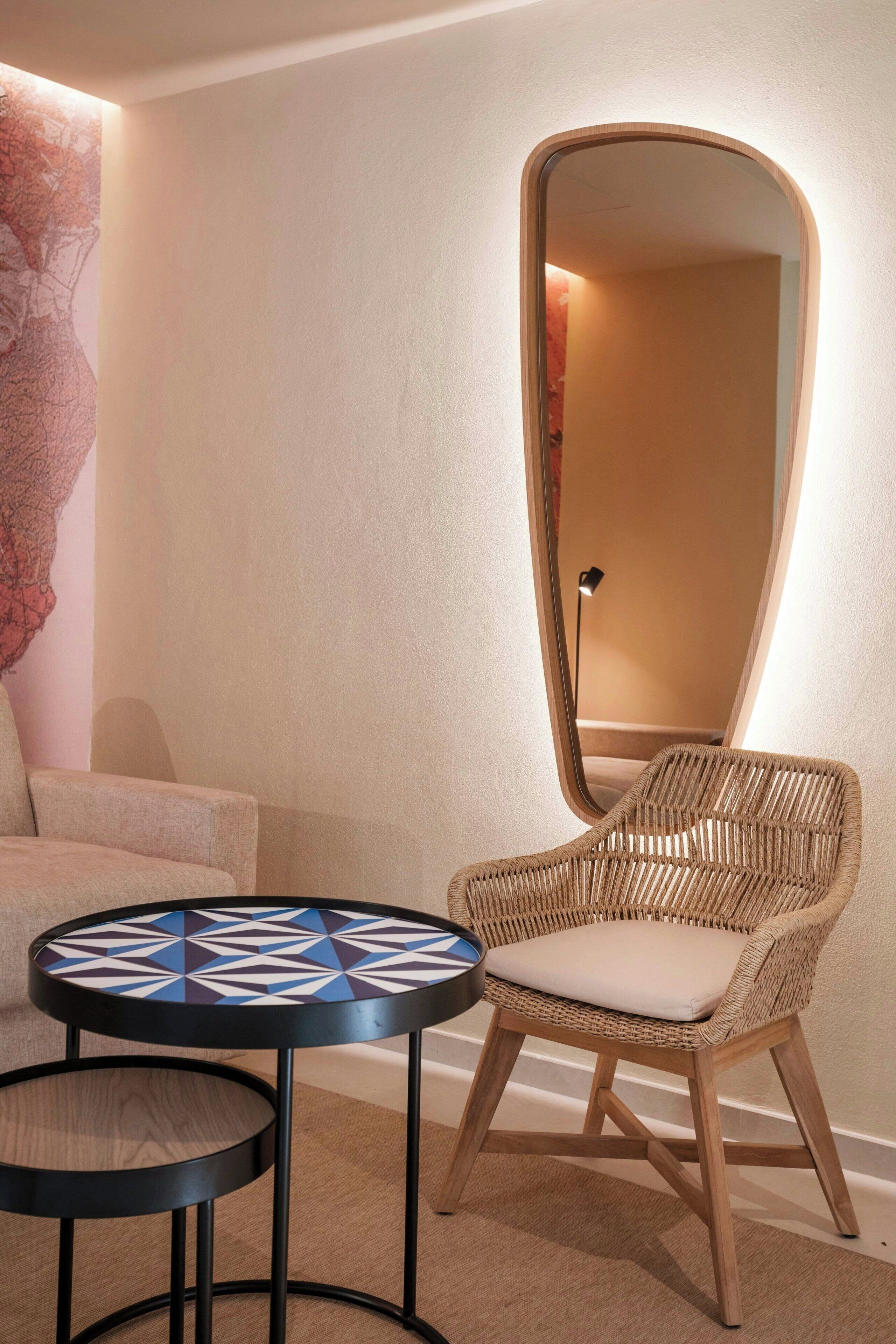 Une pièce comprenant un fauteuil en osier, une table ronde dont le plateau est orné d’un motif géométrique, et un grand miroir accroché au mur.