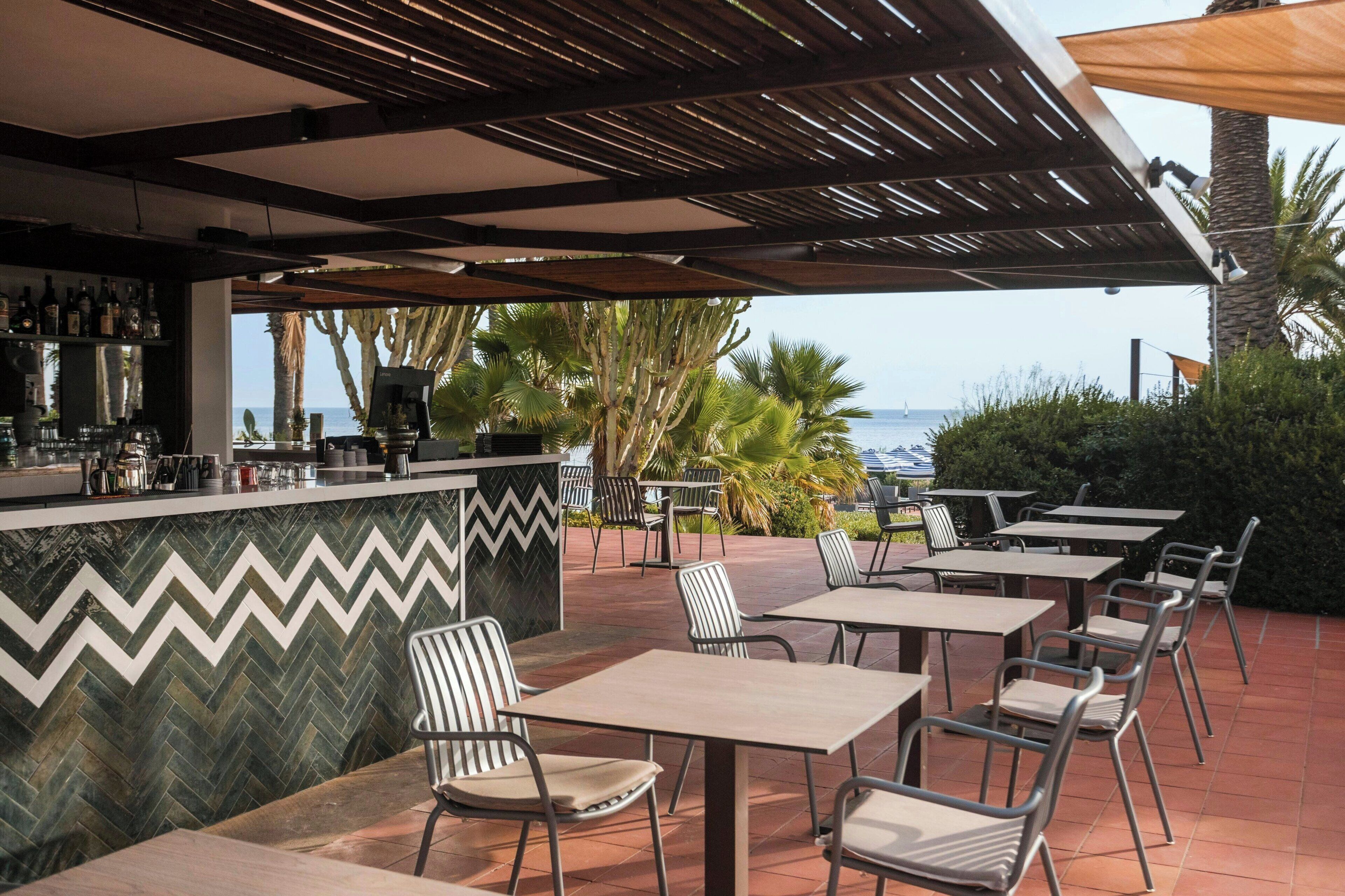An outdoor seating area with tables and chairs, a bar with a zigzag patterned wall, and a view of the sea.
