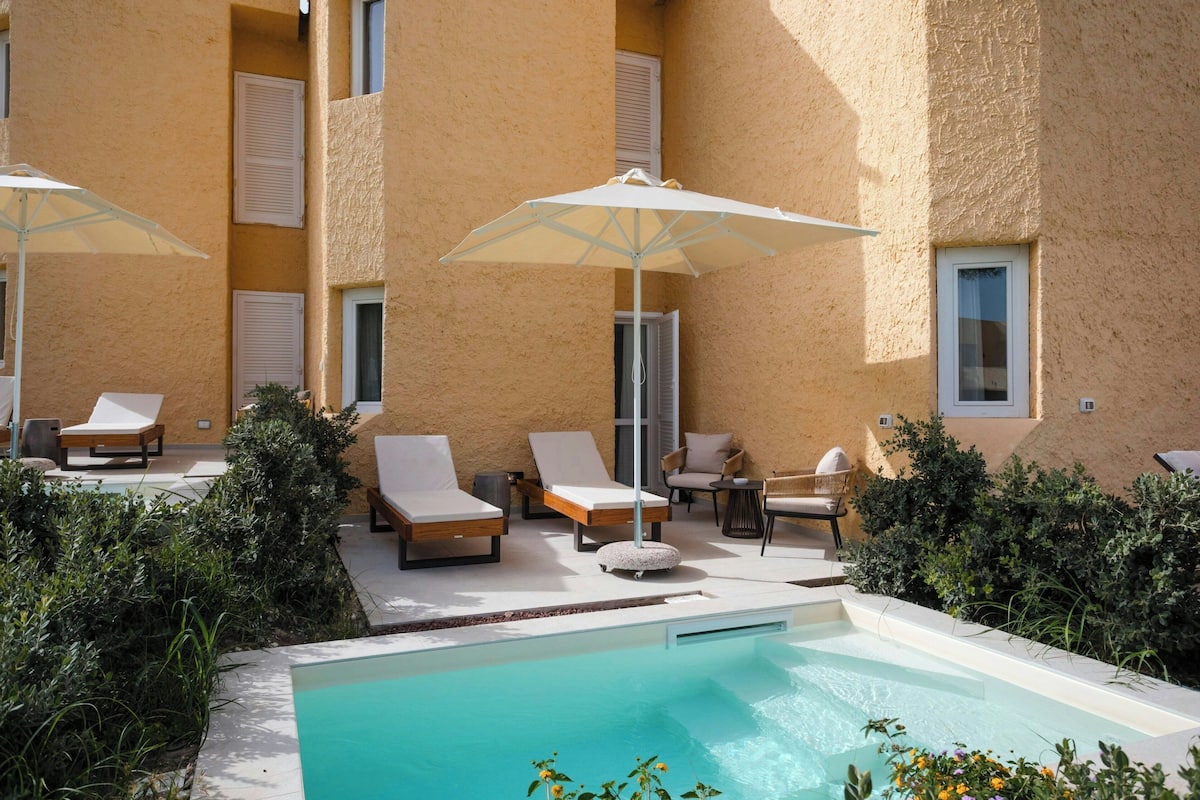 A poolside area with lounge chairs and umbrellas, surrounded by greenery and a building with beige walls and windows.