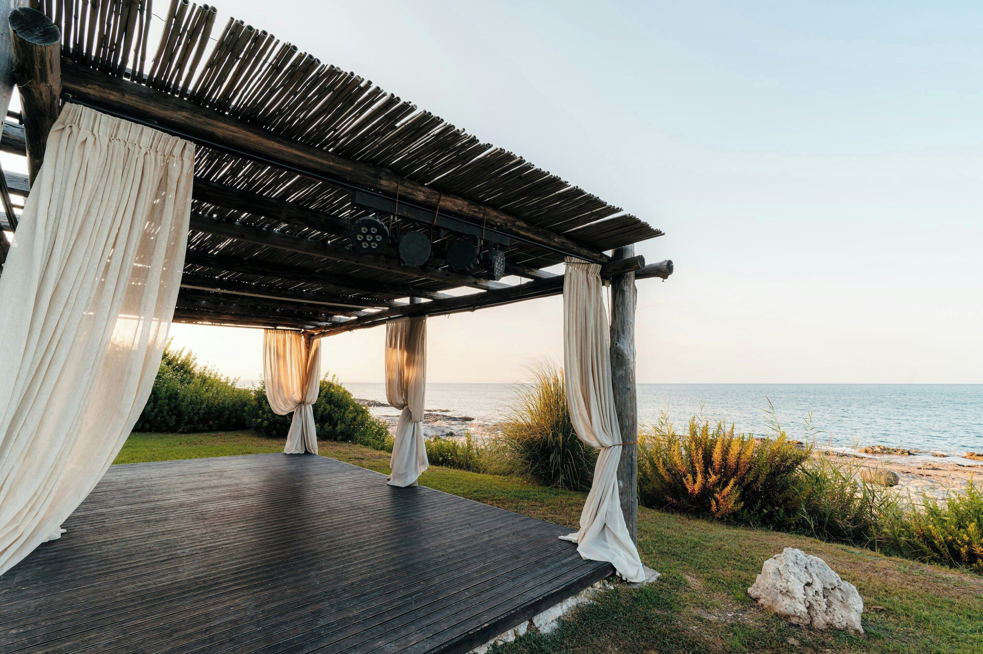 A wooden pergola with white drapes, overlooking a beach.