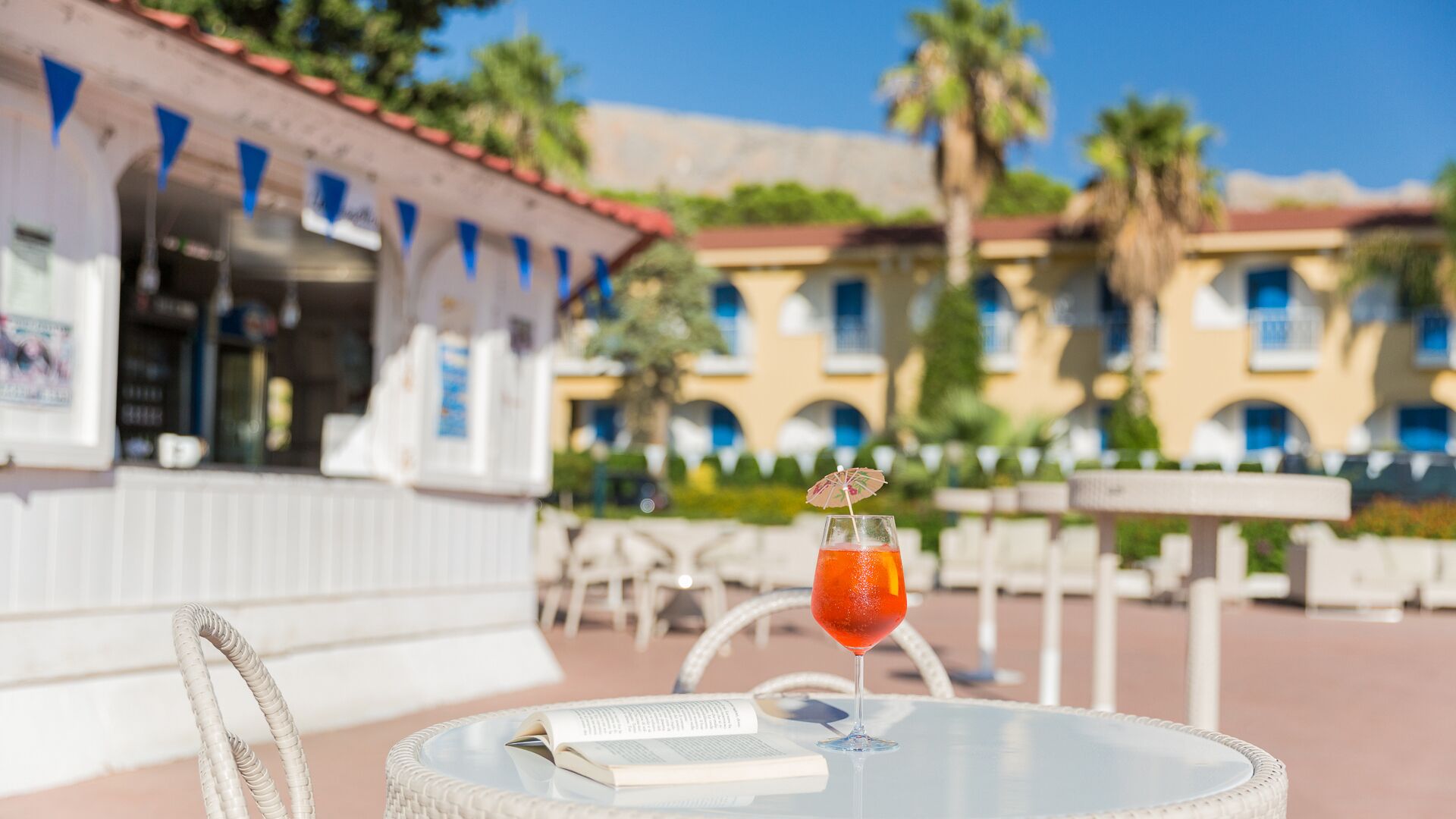 A round table with a drink and a book, set against a backdrop of a building with balconies and palm trees.