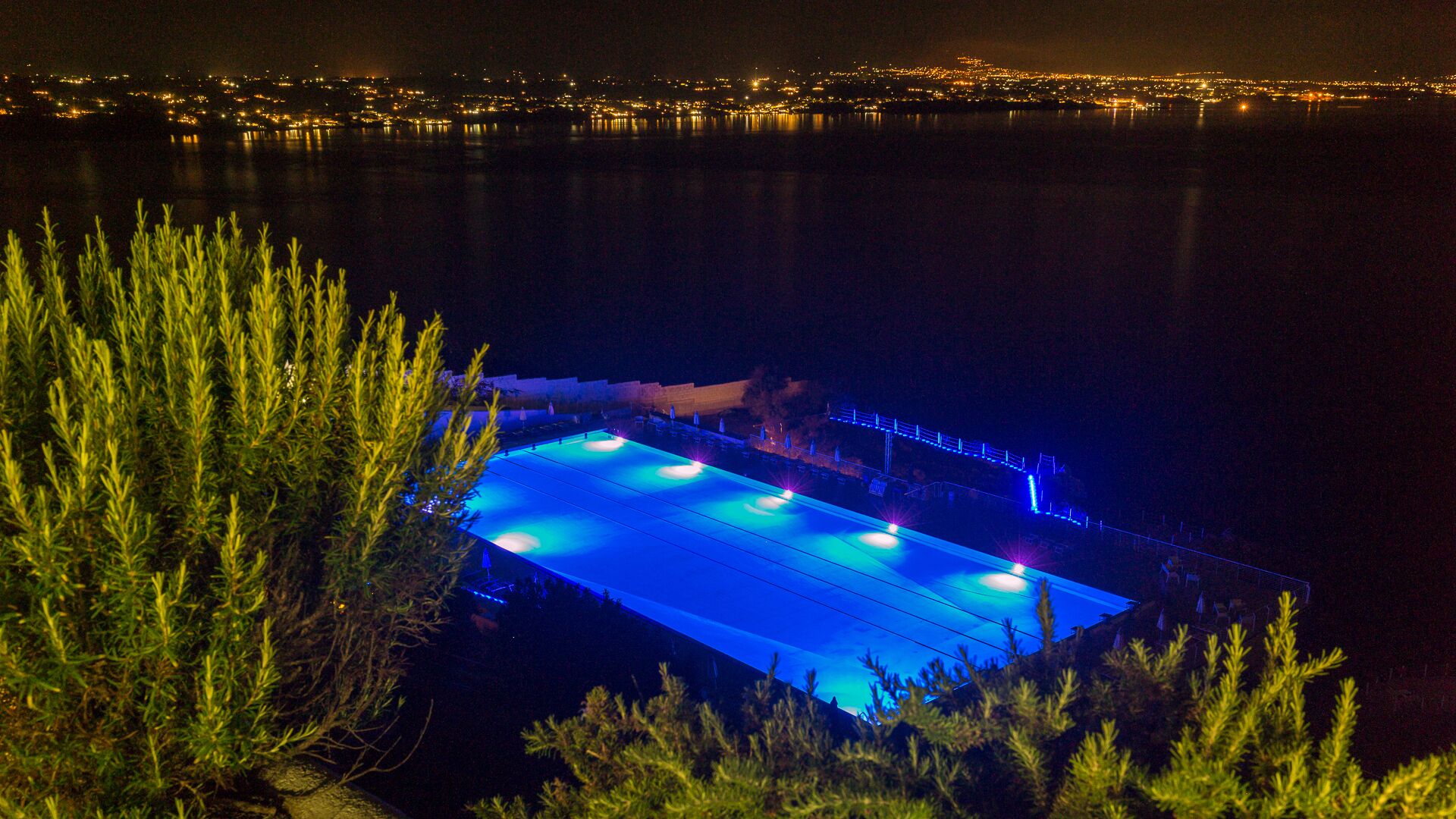 A pool with blue lighting, surrounded by greenery, overlooking a cityscape at night.