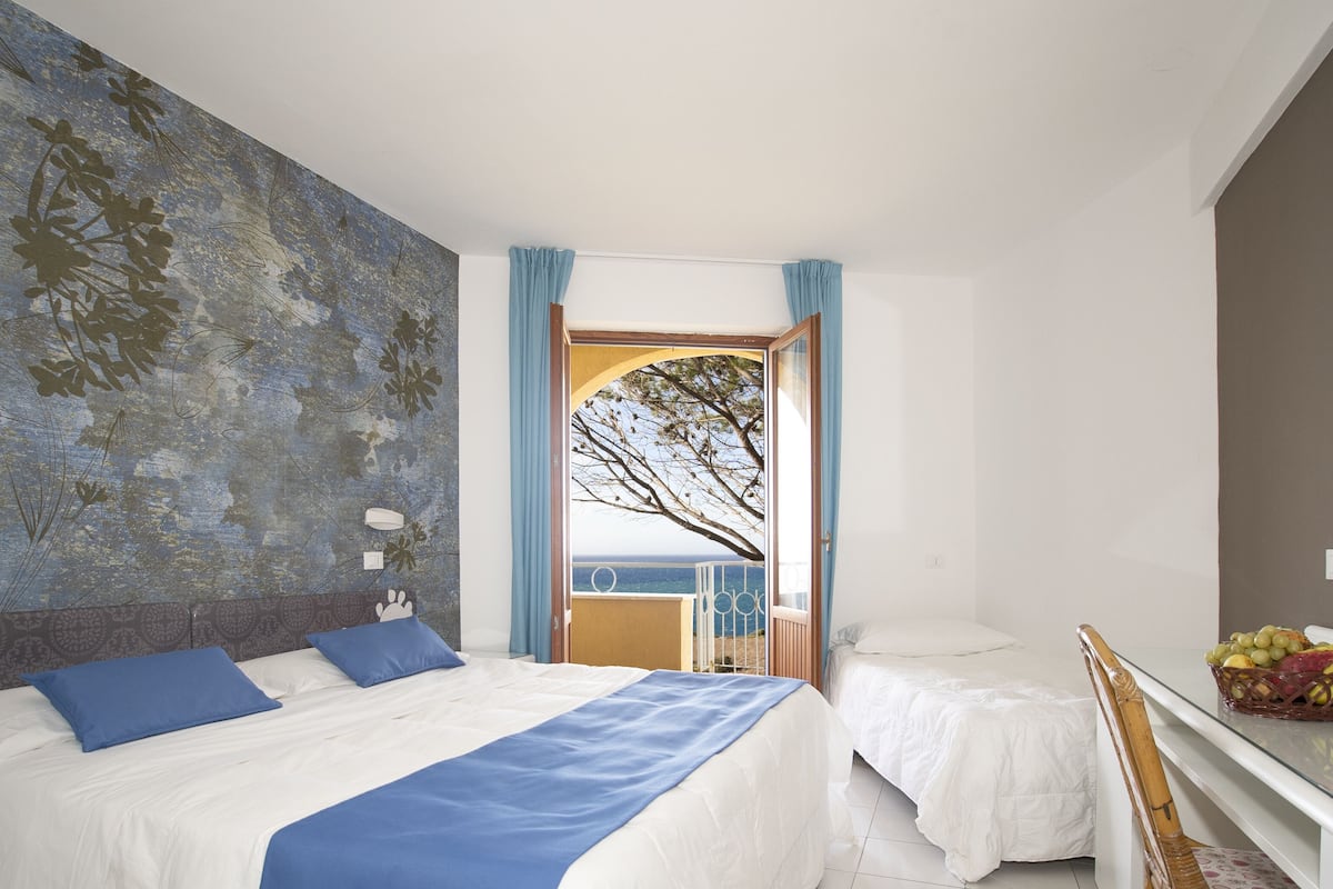 Un dormitorio con una cama grande, una cama más pequeña, una mesita de noche, un aparador, una ventana con cortinas y vista al mar.