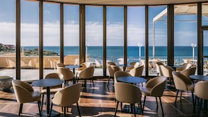 Una terraza con mesas y sillas redondas, y amplios ventanales con vistas al mar.