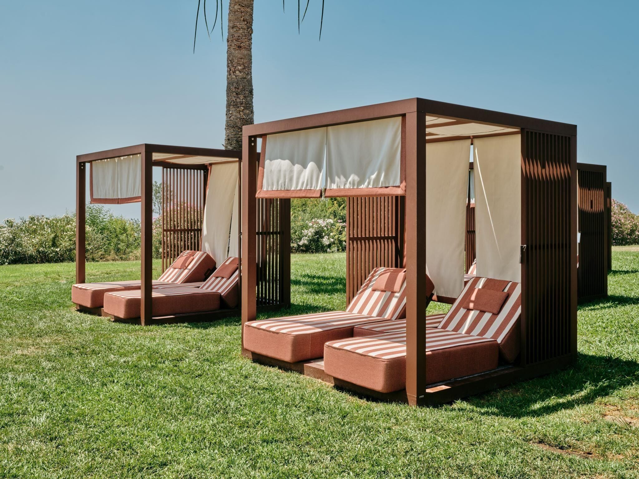 Two outdoor wooden beds with striped cushions and white curtains, set on a grassy lawn with a palm tree in the background.