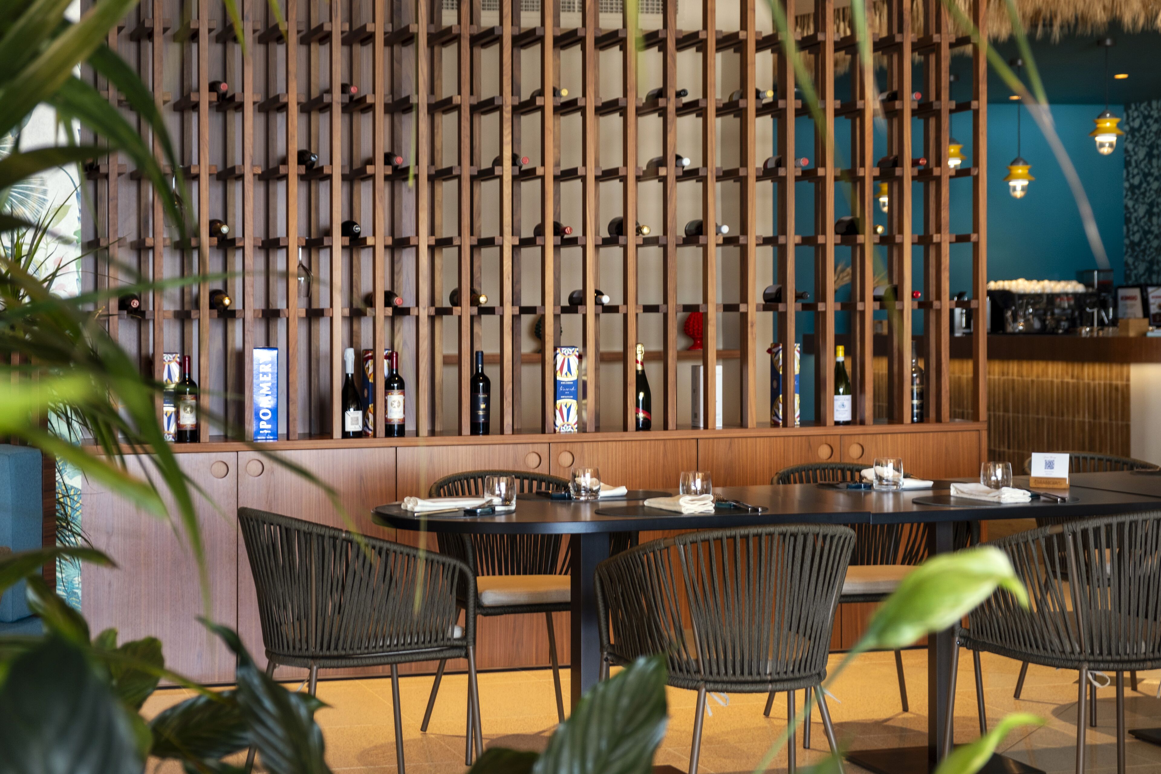 Un restaurant doté d’une longue étagère à vin en bois, de chaises en osier noir et d’un mur bleu.