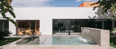 Un área de piscina moderna al aire libre con una cascada, rodeada por un edificio minimalista y abundante vegetación.